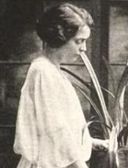 A white woman, standing, wearing a loose and drapy white dress.