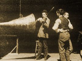 On the left is a large acoustical horn, suspended from a cord that rises out of the frame. A man plays a violin in front of it. To the right, two men dance together.