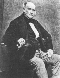 Picture of light-skinned middle-aged man, balding, seated and looking at camera, wearing mid-Victorian business suit and holding a top-hat in one hand