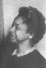 A middle-aged Black woman, smiling, wearing a dark top with a light collar, and button earrings