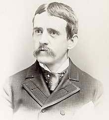 A dark-haired, mustachioed man in his early forties wearing a black coat and tie