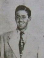 A black-haired man in a white suit and a black tie smiles as he poses for the picture