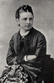 A woman with tight hair and a dark shawl top sits with her arms crossed, facing to the viewer's right with her body turned to the viewer's left