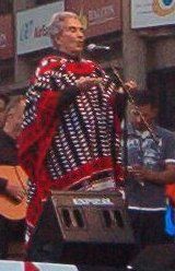 A woman standing behind a microphone on a stage; she is wearing colorful outfit and has her arms bent so that her hands are at chest-level.