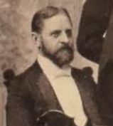 A sepia photo of a man with a mustache and beard wearing a suit with a white bowtie