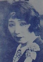 A Japanese woman with hair cut in a curled bob, wearing a jacket and tie with a corsage