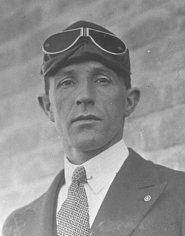 Head and shoulders image of a man wearing flying goggles, a day suit, tie and collar pin.