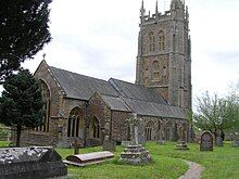 Gray stone building with ornate square tower and slate roof. In the foreground are gravestones