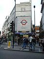 A grey-bricked building with a dark blue, rectangular sign reading "LEICESTER SQUARE STATION" in white letters all under a bright sky