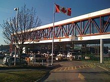 An overhead walkway is above several rows of cars waiting to cross the border