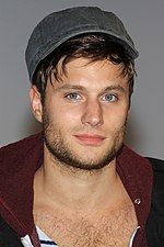 A man with blue eyes and a flatcap faces the camera. He wears a navy and white stripped top with an unzipped hoodie over the top and his chest hair on show