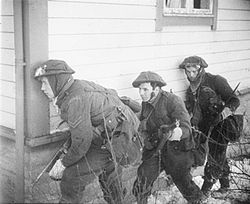 A black and white photograph of three British soldiers advancing beside a wooden house. The lead soldier is carrying a Thompson submachine gun and is peering around the corner of the house, while the other two are carrying rifles.