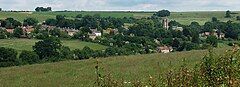 The roofs of many houses, and a church spire can be seen in a green valley with several trees.