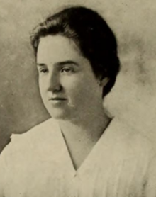 A young white woman with dark hair dressed back to the nape, wearing a white blouse (might be an academic gown)