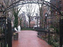 A color photograph of Christopher Park in winter, showing the wrought iron entrance arch in the foreground and the brick pavement surrounded by five and six-story brick buildings; in the center background are four white statue figures: two males standing, one with his hand on the other's shoulder and two females seated on a park bench, one woman with her hand touching the other's thigh. All are dressed in jeans and loose clothing