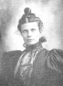 Emma S. Yule, a young white woman with dark hair in a updo, wearing a dark dress with large puffy sleeves and lace inset yoke