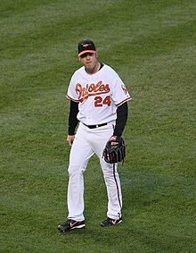 A man in a white baseball uniform with orange script across the chest and a black baseball cap walks across a grass field with a black baseball glove on his left hand