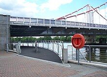 Start of a footbridge that curves under one end of a suspension bridge, along the bank of a wide river.