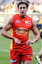 A male athlete with light brown hair wearing a sleeveless jersey and shorts walks on the grass surface of the playing arena.