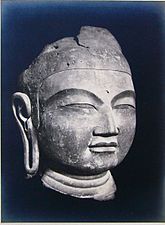 A Buddha head in three-quarter view with elongated earlobe, thick lips, narrow slit eyes and broad nose. Black and white picture.