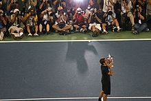 A man in black shorts and a black short kisses a silver trophy, with dozens of photographers shooting him in the background