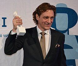 Ville Virtanen wearing a white shirt and dark blazer, standing onstage holding a trophy, grinning and looking left of camera