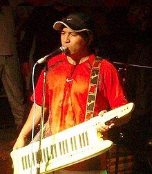 Pablo Lescano wearing a bright-colored t-shirt with the Nike swoosh logo as well as a black Nike ball cap, playing a keytar onstage, appearing to sing or speak into a microphone