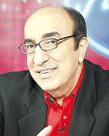 Elias Rahbani wearing glasses, red button-down shirt, and black blazer, smiling at camera with head tilted and microphone pinned to shirt