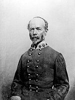 Black and white photograph shows a balding man with a salt-and-pepper moustache and beard. He wears a double-breasted gray military uniform with three stars on the collar.