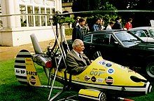 Small, one man, open-cockpit helicopter on a lawn about the size of a car next to it, with a man sitting in it.