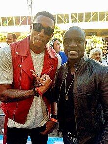 Two black men pose for the camera. The left man is taller and bends slightly to the right in order to fit into view. He wears black sunglasses, a white t-shirt with a red vest, and black pants. He has his right hand clasped in a gesture with the right hand of the man on the right, who makes a peace symbol gesture with two fingers of his right hand. The man on the right wears a black leather jacket over a black shirt, a silver-colored necklace, and blue jeans with a studded belt.