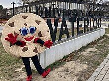 A cookie mascot stands in front of a sign that reads University of Waterloo and the Dana Porter Library