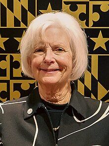 Klausmeier smiling in front of the seal of Baltimore County, wearing a black blazer jacket, black shirt, and gold necklace.