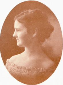 A young white woman with dark hair, in profile, wearing a light-colored dress with an off-the-shoulders smocked neckline; in an oval frame