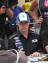 A man in his late twenties wearing a black T-shirt with sponsors logos and a blue and white baseball cap on his head. He is looking to the right and is holding a black pen in his right hand