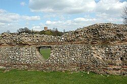 Mortared wall with stacked thick stone layers over thin red brick layers, with a triangular tunnel through