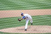 A man in a white baseball uniform and a blue baseball cap throws a baseball from a dirt mound on a grass field. He is throwing with his left hand, and there is a black baseball glove on his right hand.