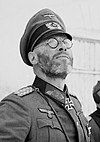 A black-and-white photograph of a man wearing a military uniform and a neck order in shape of an Iron Cross. He is wearing glasses and is looking up into the sky.