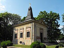 A small, yellow neoclassicist building with a tower resembling a victory column or tombstone, topped with a golden cross. Stairs lead up to the door under a triangle-shaped tympanum.It has three windows on it visible side, all surrounded with light pink marble and fenced. It is surrounded with a park.