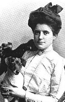 A young white woman with dark hair in a bouffant updo, wearing a white high-necked blouse with a cameo pin, holding a small short-haired dog.