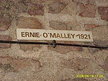 "Colour photograph of the a brick wall with a cream plaque bearing the words "Ernie O'Malley 1921" in dark-brown capital letters"