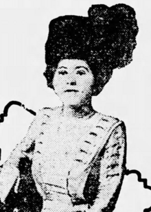 A white woman with dark hair, wearing a large dark asymmetrical hat and a light-colored dress