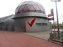 An outside view of the Benfica Museum. Its façade consists of grey metal panels with a red wing- or eagle-shaped piece of art followed by "MUSEU BENFICA COSME DAMIÃO" in black letters. On the top of the building, there is a red geodesic dome with a grey covering.