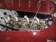 Close-up of a straight-eight engine installed in the front of a racing car, as seen through the right side of the bonnet, which is hinged in the centre and is held open with a metal rod. There are four downdraft carburettors on the near side of the engine, these have intake stacks angled backward and toward the engine. The exhaust manifold is visible on the same side of the engine as the carburettors.