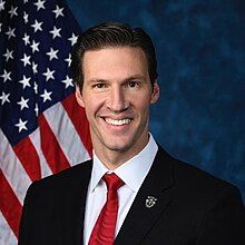 Official House portrait of Harrigan smiling in front of the U.S. flag, wearing a black suit with military medallion, white shirt, and red tie.