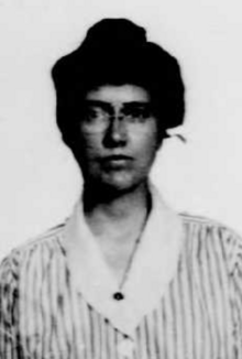 A white woman, with dark hair in a bouffant updo, wearing a striped top with a white collar, and glasses