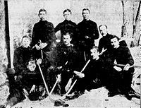 Men wearing hockey sweaters assembled in three rows in front of a painting of trees. Several are holding ice hockey sticks