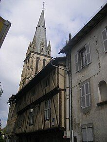 A tall stone church tower with a pointed roof rises above a weathered, half-timbered building with a gray facade.