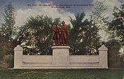 Picture postcard of a large bronze statue of two men. The statue is on top of a stone pedestal that is flanked on each side by stone walls with stone benches running along the base of the wall. The pedestal, walls, and benches sit on top of a large stone step. The monument has grass in front of it, and trees behind it. There is printing along the top of the postcard that says "No. 140. Goethe–Schiller Monument. Washington Park. Milwaukee, Wis."