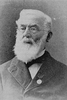 A black-and-white photographic portrait of a bearded, white-haired bespectacled man, shown from the chest up